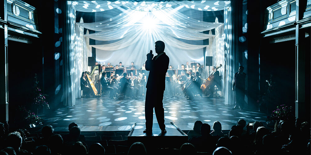 Theaterball_2025_D.Rückschloss Festlich beleuchteter Theaterraum mit einer Person auf einem Laufsteg vor einer Bühne. Im Hintergrund sitzt ein Orchester, während das Publikum im Zuschauerraum zu sehen ist.
